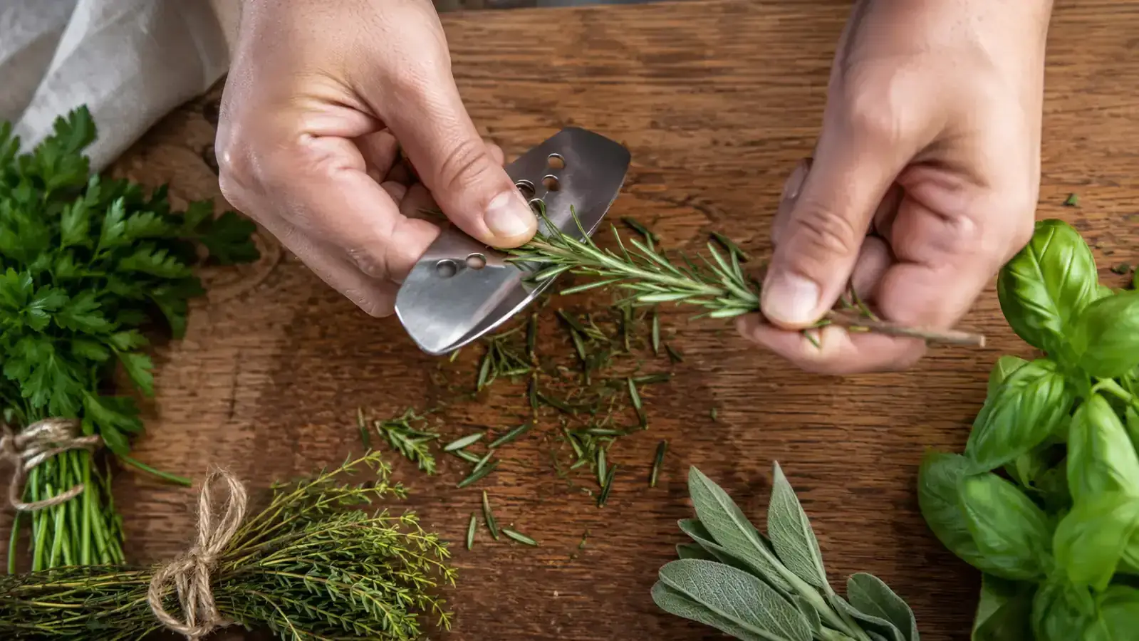 Hands stripping fresh rosemary and thyme with a stainless herb stripper over a wooden cutting board