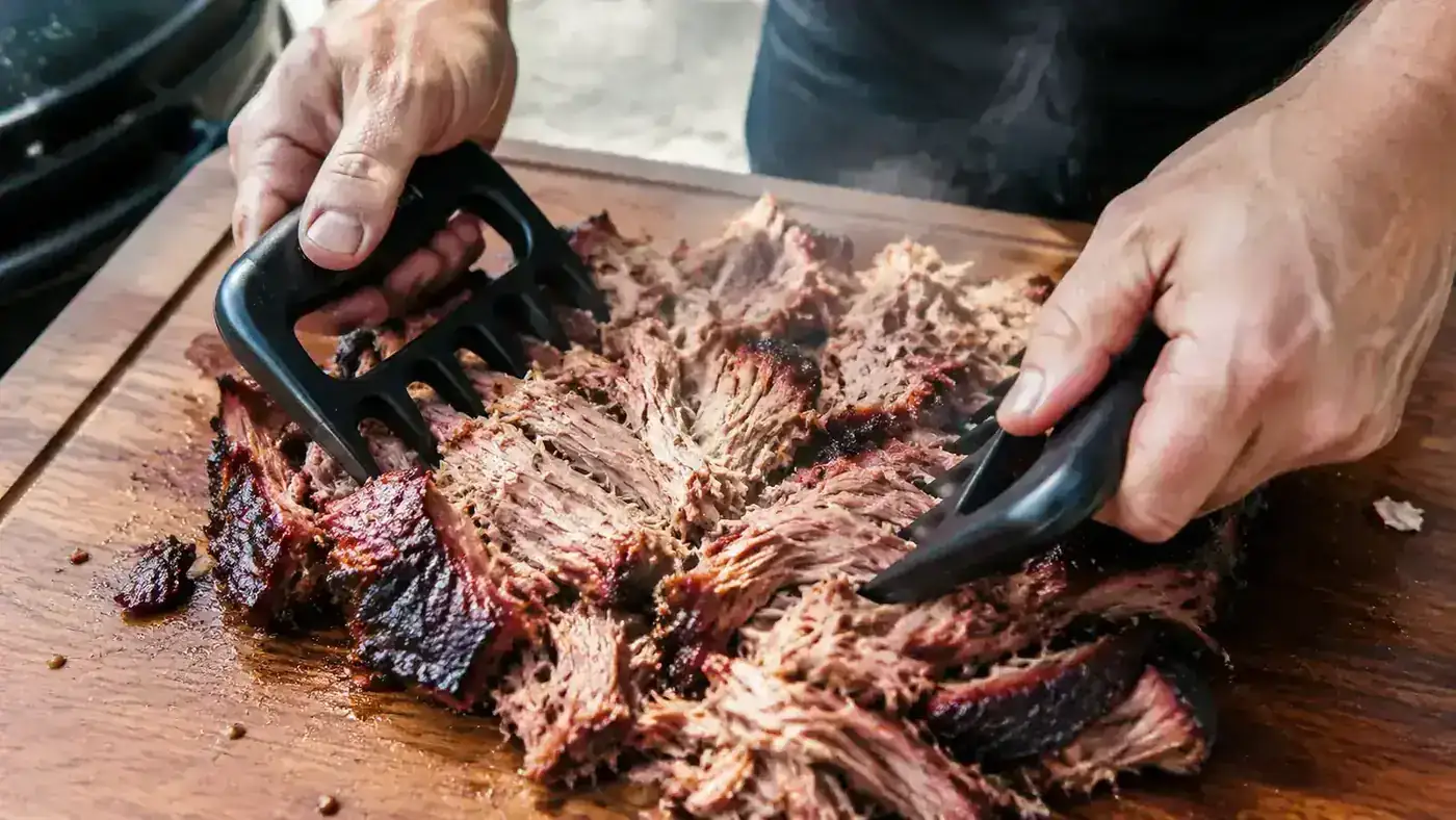 Two hands using stainless-tipped meat claws to shred a whole pork shoulder on a cutting board