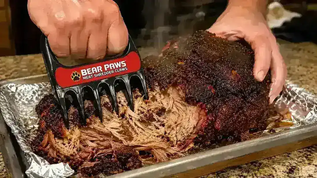 Hand wearing a meat claw shredding a pork shoulder straight out of the smoker on a sheet pan