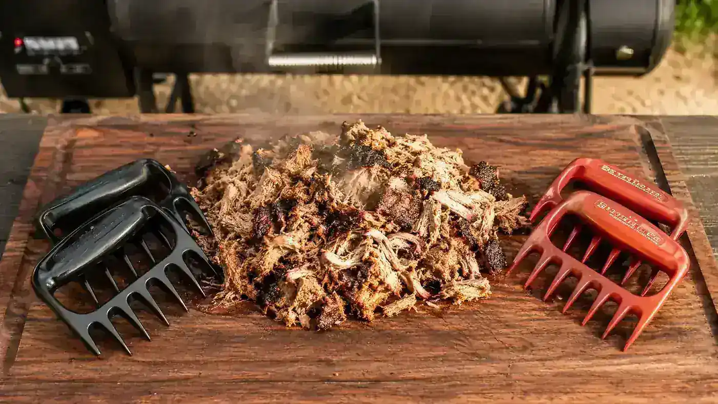 Two pairs of meat shredder claws crossed over a cutting board next to a pile of freshly pulled pork shoulder