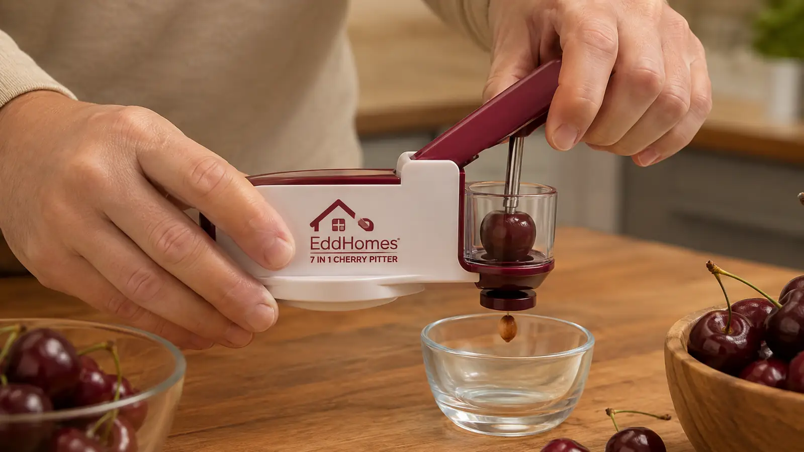 A hand squeezing an EddHomes cherry pitter over a bowl, pit dropping into a smaller catch bowl below