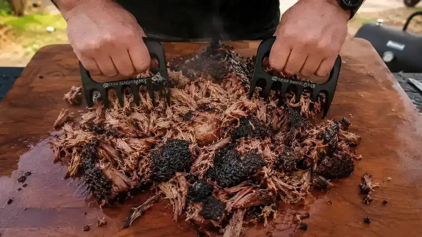 Hands using stainless-tipped meat claws to shred a smoked pork shoulder on a wooden cutting board