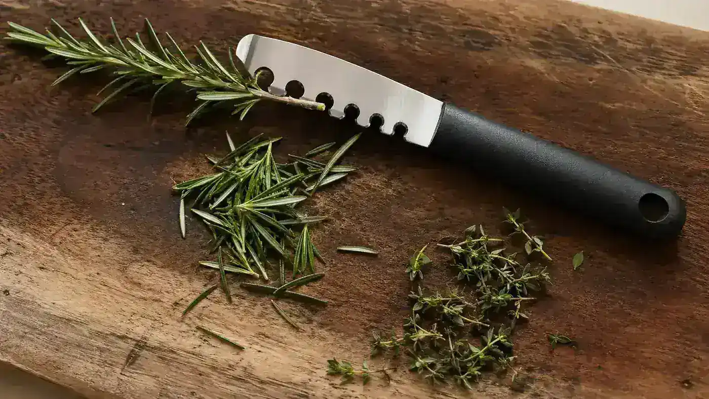Stainless steel herb stripper with nine holes lying next to a small pile of stripped rosemary and thyme leaves on a wooden cutting board